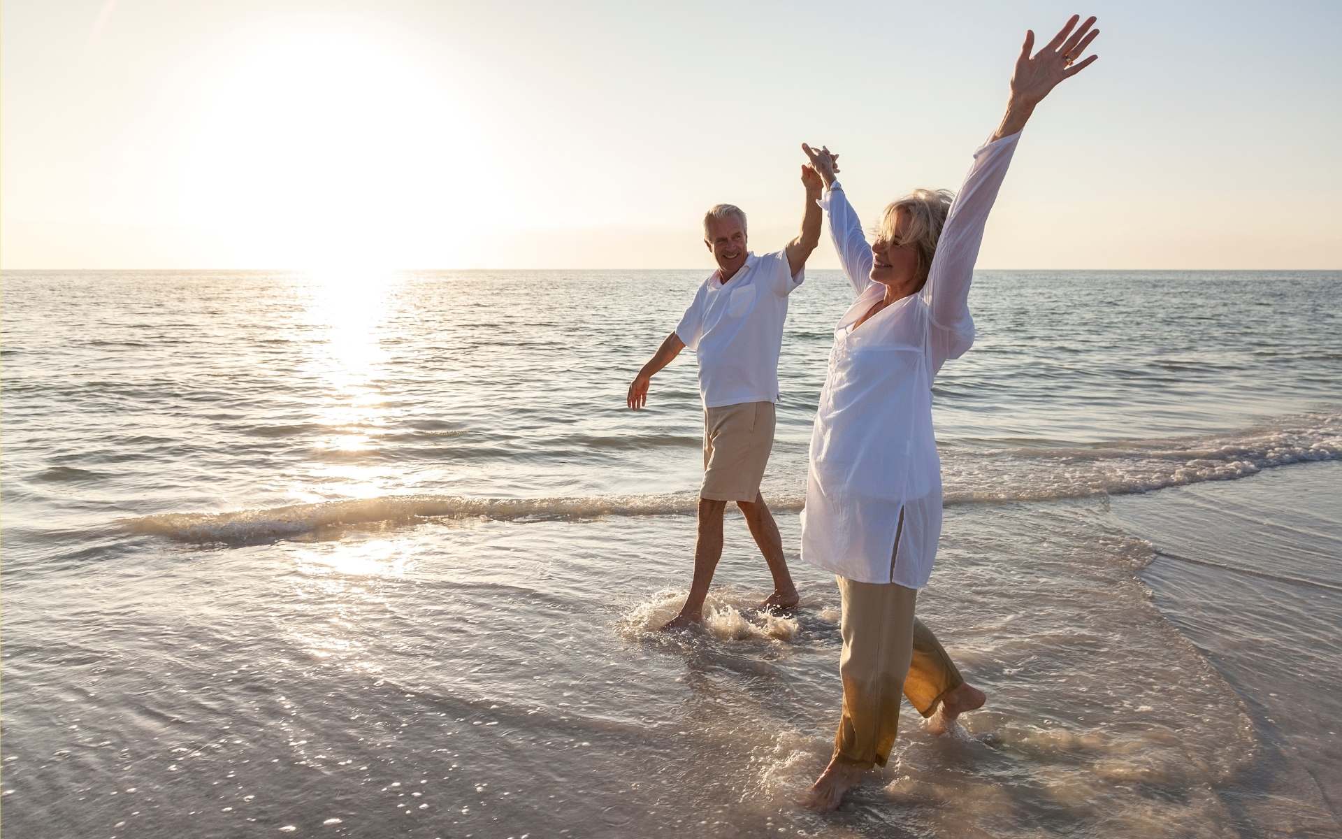 Glückliches Paar läuft mit gehaltenen, in den Himmel gestreckten Armen am Strand entlang und genießt die Sonnenstrahlen. Beide in weiß gekleidet. 